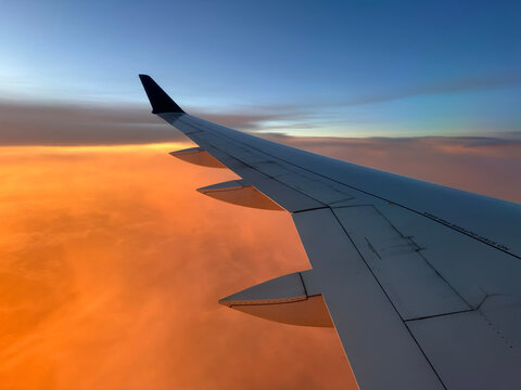 View from an aircraft window of a passenger airplane wing with a dark winglet and flaps extended. A layer of orange clouds lies below the wing at sunset, with a blue gradient sky above.