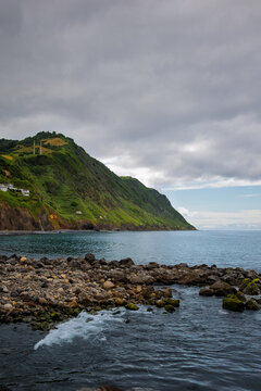 Povoa&ccedil;&atilde;o coastline showing volcanic rocky shore with ocean water, lush green hills covered in terraced fields, and a cloudy sky above the Atlantic Ocean