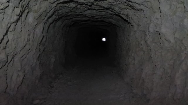POV walking through a dark narrow cave tunnel in a rock face toward the bright light at the end, exploration concept.