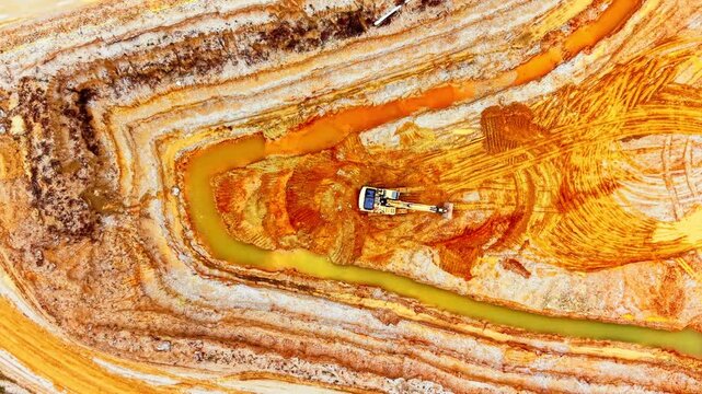 Close aerial view of excavator in sand quarry. Machine digs beside a winding yellow stream cutting through layered ochre and rust colored sand.