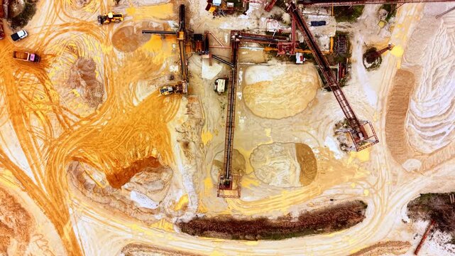 Front loader moves sand near quarry conveyor belt. Overhead view shows stained sand mounds, steel towers and a truck parked beside the pit.