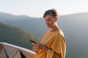 Obraz premium Middle aged woman using smartphone on a mountain balcony at sunrise for lifestyle background and relaxation scene