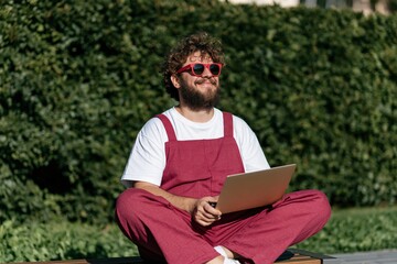 Fototapeta premium Curly haired man wearing red overalls using a laptop outdoors on a bench for remote work background and lifestyle banner design