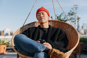 Obraz premium Rooftop relaxing man in red beanie sitting in a hanging wicker chair with coffee mug for lifestyle background and urban leisure design
