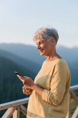 Naklejka premium Older woman holding phone and drink on balcony overlooking mountains for travel background and wellness lifestyle banner