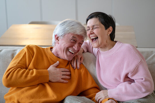 Happy senior married couple laughs and embraces on sofa, cheerful wife hugs smiling husband, affectionate elderly spouses share humor, emotional closeness and positive retirement relationship.