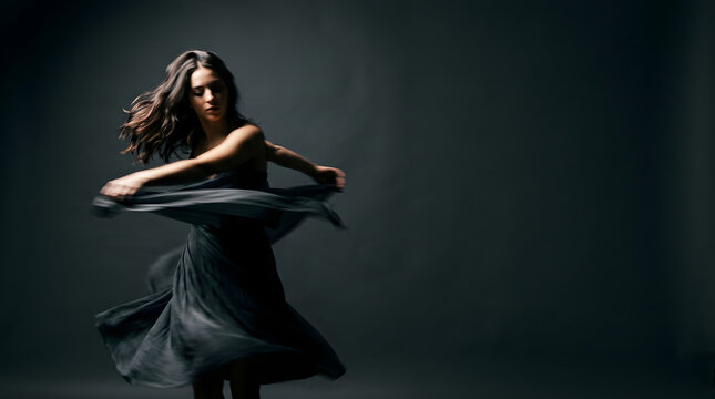 Elegant young woman dancing in a flowing dark dress against a moody studio background with motion blur effect.