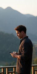 Obraz premium Young man on mountain terrace reading smartphone with drink at dusk for lifestyle background and travel banner