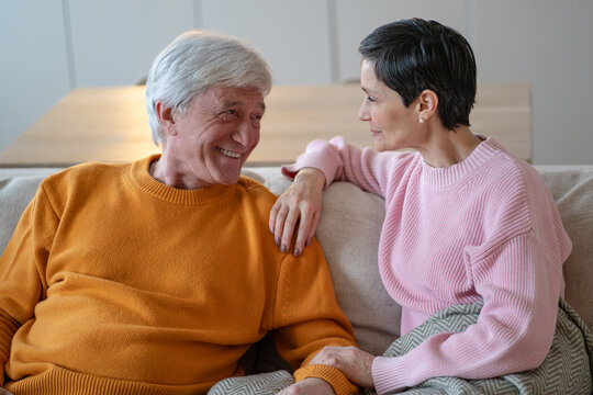 Affectionate elderly husband and wife sit close on couch, smile at each other as woman holds hands with partner, senior married couple shares loving conversation, trust and emotional connection.