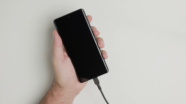 Male hands holding smartphone and plugging charger cable then unplugging on white background, battery charging concept, top view, vertical view, slow motion