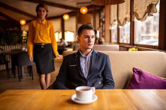 Young man sitting alone at a restaurant table with a woman in a yellow short on background