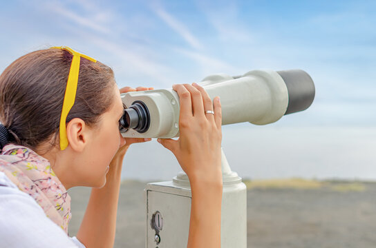 A young woman looks through a public viewer telescope against a blue sky background.