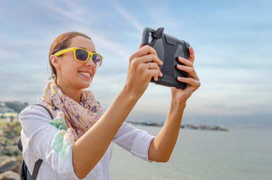 A smiling young woman wearing yellow sunglasses takes a photo with a digital tablet outdoors near the water.