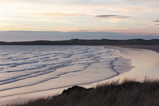Rolling waves and parallel surf lines on a sandy beach at sunset with dune grasses in the foreground, Newborough, Anglesey, North Wales