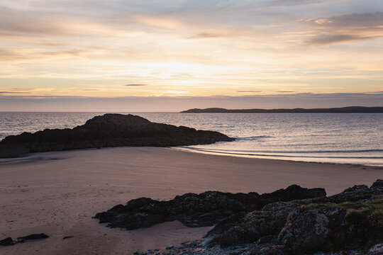 Rocky outcrop silhouetted against a pastel sunset sky on a sandy beach with distant headland, Llanddwyn Island, Anglesey, North Wales