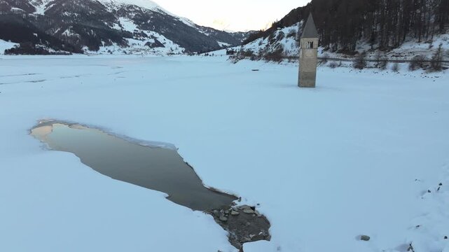 Aerial view of the historic submerged bell tower rising from the frozen Reschensee lake surrounded by snow-covered mountains in Graun im Vinschgau, Trentino-Alto AdigeS, Italy.