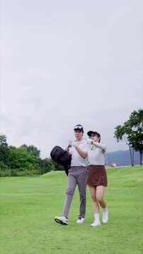 Asian couple walking together across green golf course carrying golf bag under overcast sky