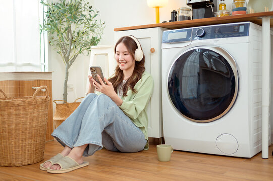 Asian Woman Using Smartphone and Listening to Music While Doing Laundry