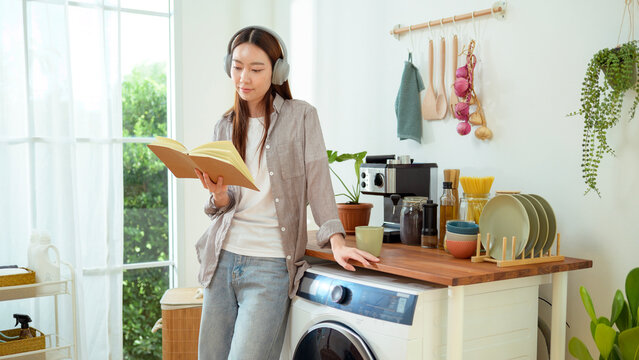 Young Woman Reads Book While Standing Next to Washing Machine in Home Kitchen