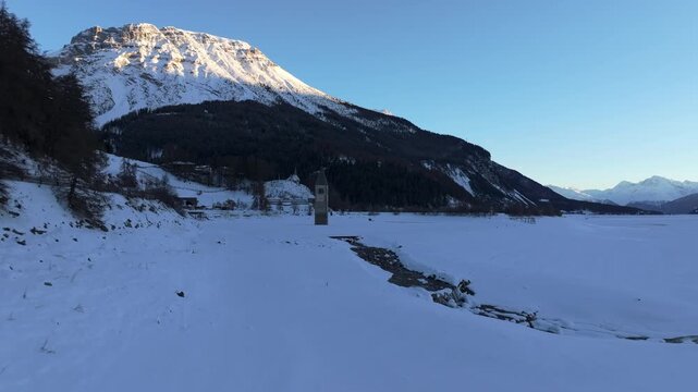 Aerial view of the historic sunken bell tower of Curon rising from the frozen surface of Lake Reschen surrounded by snow-covered mountains Graun im Vinschgau, Trentino-Alto AdigeS, Italy.