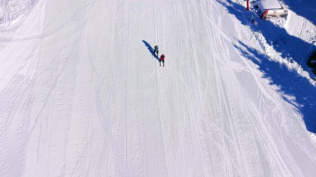 Two skiers pausing on sunlit piste near chairlift station. Overhead view captures long shadows on groomed snow beside a wooden lift shelter and equipment shed.