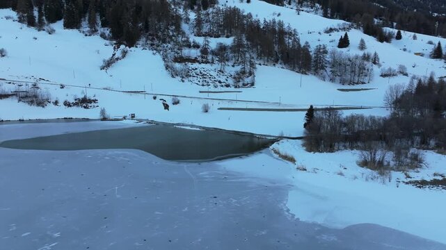 Aerial view of the frozen Reschensee lake surface surrounded by snow-covered mountains and pine forests in Graun im Vinschgau, Trentino-Alto Adige, Italy.