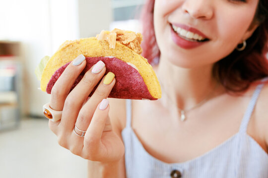 Food blogger lifestyle shot: smiling young woman holding a double-shell taco with colorful manicure. Snack enjoyment concept.