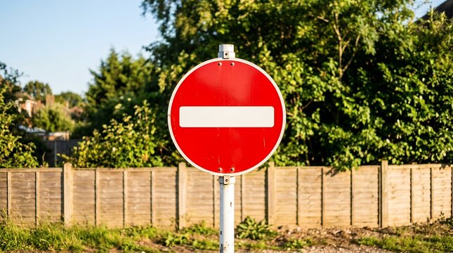 Red no entry road sign outdoors, circular stop traffic symbol, metal prohibition street sign with white bar against green trees and fence background