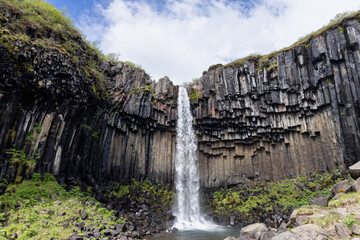 Svartifoss waterfall drops into a rocky pool beneath a broad wall of dark basalt columns in Skaftafell, Iceland. Green moss and boulders frame the falls under a bright, cloudy sky. © Artem