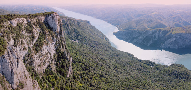 Sheer vertical limestone pinnacle rises dramatically beside a turquoise Danube river winding through a vast green canyon of Iron Gates. Adventure concept.