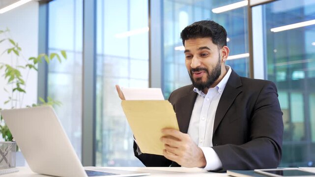 Surprised smiling businessman reading letter with great news sitting at workplace in business office. Excited worker in formal suit is celebrating success, happy to receive positive good notification.
