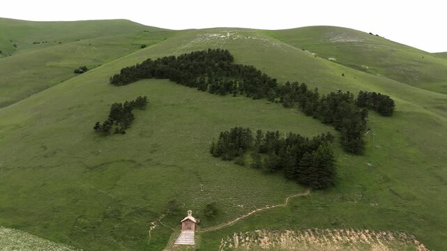 Aerial view of the Italy-shaped forest plantation on a green hillside with a small chapel in Castelluccio, Umbria, Italy.