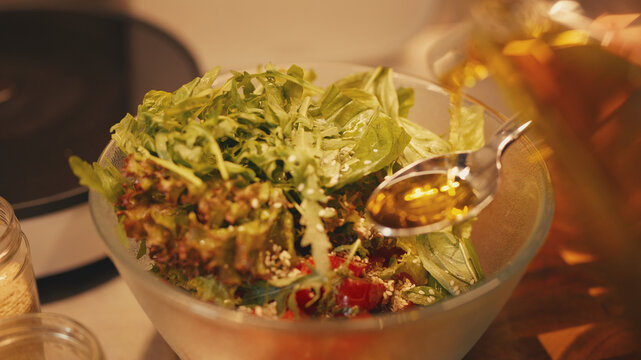 Close-up of a person pouring olive oil from a glass bottle into a tablespoon over a fresh vegetable salad. Dressing salad with tomatoes and greens in a glass bowl. Healthy home cooking.