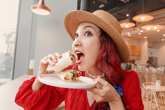 Young woman in straw hat and red dress enjoying a fresh wrap burrito for lunch at a bright modern cafe, smiling and relaxed
