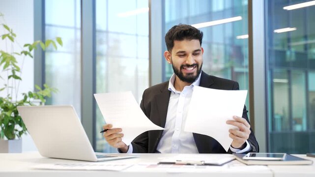 Happy financier satisfied with financial results sitting at workplace in business office with computer. Smiling businessman investor in formal suit looks at documents, celebrates successful investment
