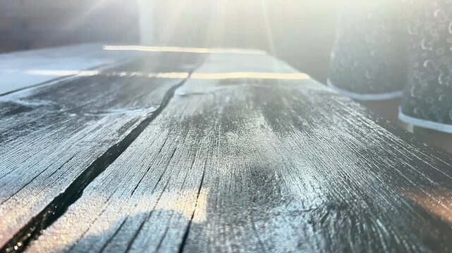 Close-up of a paintbrush applying dark protective wood stain or paint onto a rustic wooden table surface outdoors in bright backlighting.
