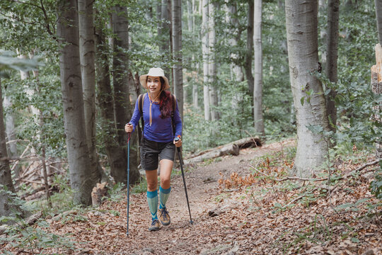 Smiling woman with trekking poles and vivid compression socks strides confidently down a sunlit beech forest trail. Adventure concept.