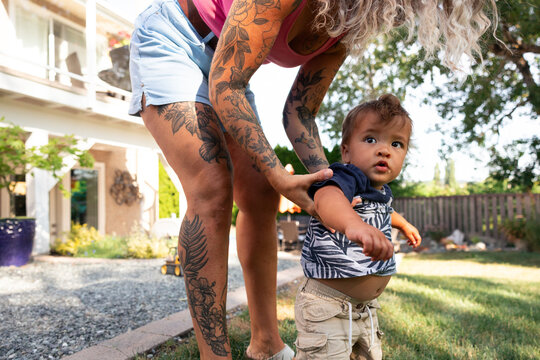 Candid mother with tattoos helping toddler walk in backyard