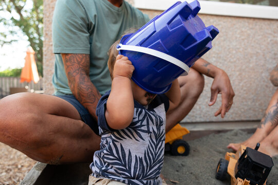 Toddler playing with blue bucket in sandbox with father nearby
