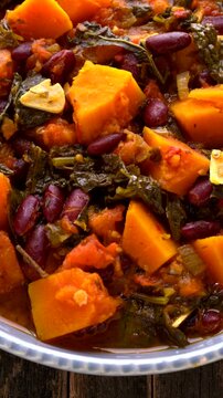Vertical close-up of a healthy vegan stew with Hokkaido pumpkin, kale, and red kidney beans. Slow spin of cooked cavolo nero kale goulash in a blue bowl on a dark, rustic table. Vertical video.