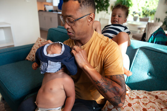 Candid father dressing baby at home with playful toddler nearby