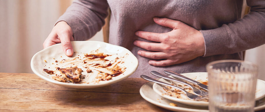 Person holding a dirty empty plate with hand on stomach feeling discomfort after eating meal and overeating at home with dirty dishes