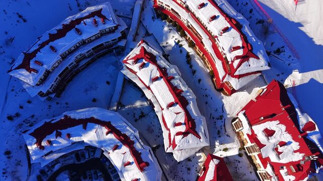 Curved buildings under snowy red roofs, Pamporovo, Bulgaria. Rows of curved hotel blocks with chimneys, heavy roof snow, a parking area with cars and a ski slope with red netting.