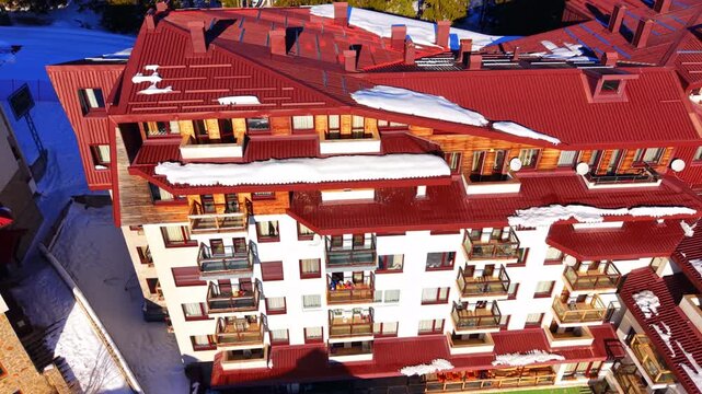 Apartment building with red roof and balconies, Pamporovo, Bulgaria. White facade, wooden cladding, glass balconies, satellite dishes, chimneys and patches of snow on the roof.