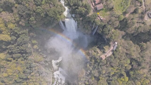 Aerial view of the cascading Marmore Falls surrounded by lush forest and a vibrant rainbow in Terni, Umbria, Italy.