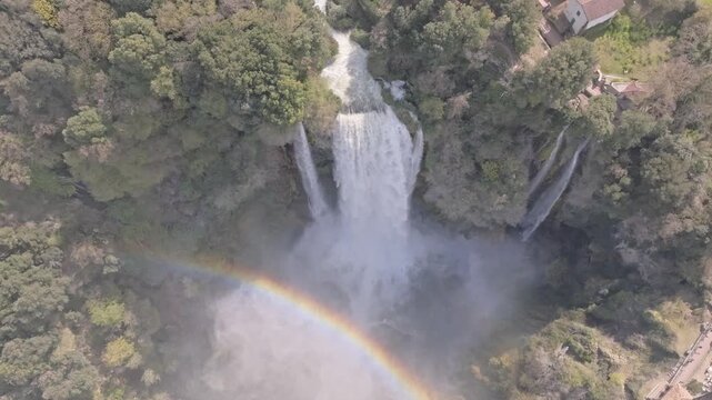 Aerial view of Marmore Falls cascading down lush green cliffs with a vibrant rainbow visible in the mist in Terni, Umbria, Italy.