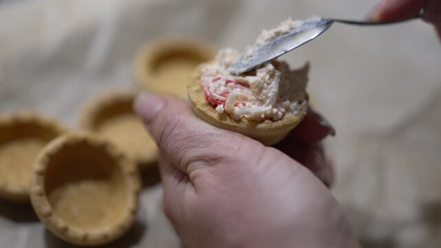 Crab salad tartlets preparation for festive appetizers. Close up shot captures hands spooning savory filling into golden pastry shells with precision.