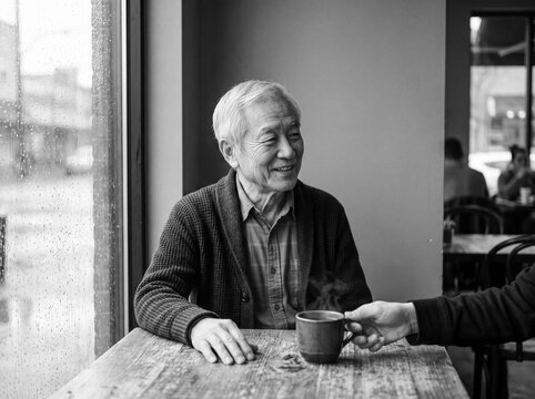 Happy elderly Asian man smiling while receiving a hot cup of coffee at a cozy cafe on a rainy day