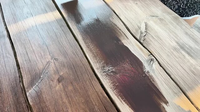 High-angle shot of a person in black rubber gloves using a paintbrush to apply dark wood stain onto rustic wooden table planks outdoors.