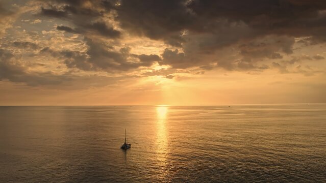 Aerial view of a lone sailboat drifting on the calm Andaman Sea during a golden sunset with dramatic clouds in Phuket, Phuket, Thailand.